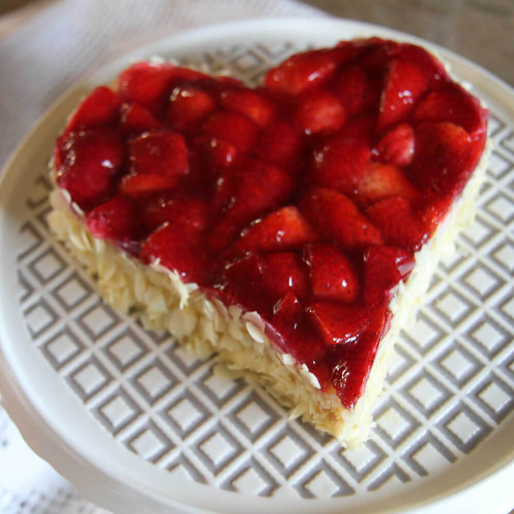 Backrahmen Herz Edelstahl Es ist ein Kuchen in Herzform zu sehen. An der Seite des Kuchens befinden sich Mandelsplitter und auf dem Kuchen Erdbeeren. Der Kuchen steht auf einen karierten Tortenplatte