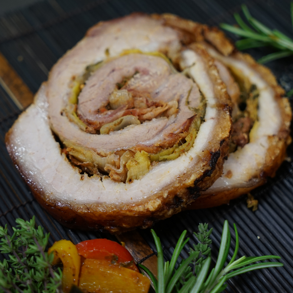 Rezept für Porchetta gefüllter Schweinebauch mit schöner Kruste, gefüllt mit Schinken und Käse auf einem schwarzen Teller mit etwas Grillgemüse und frischen Kräutern serviert.