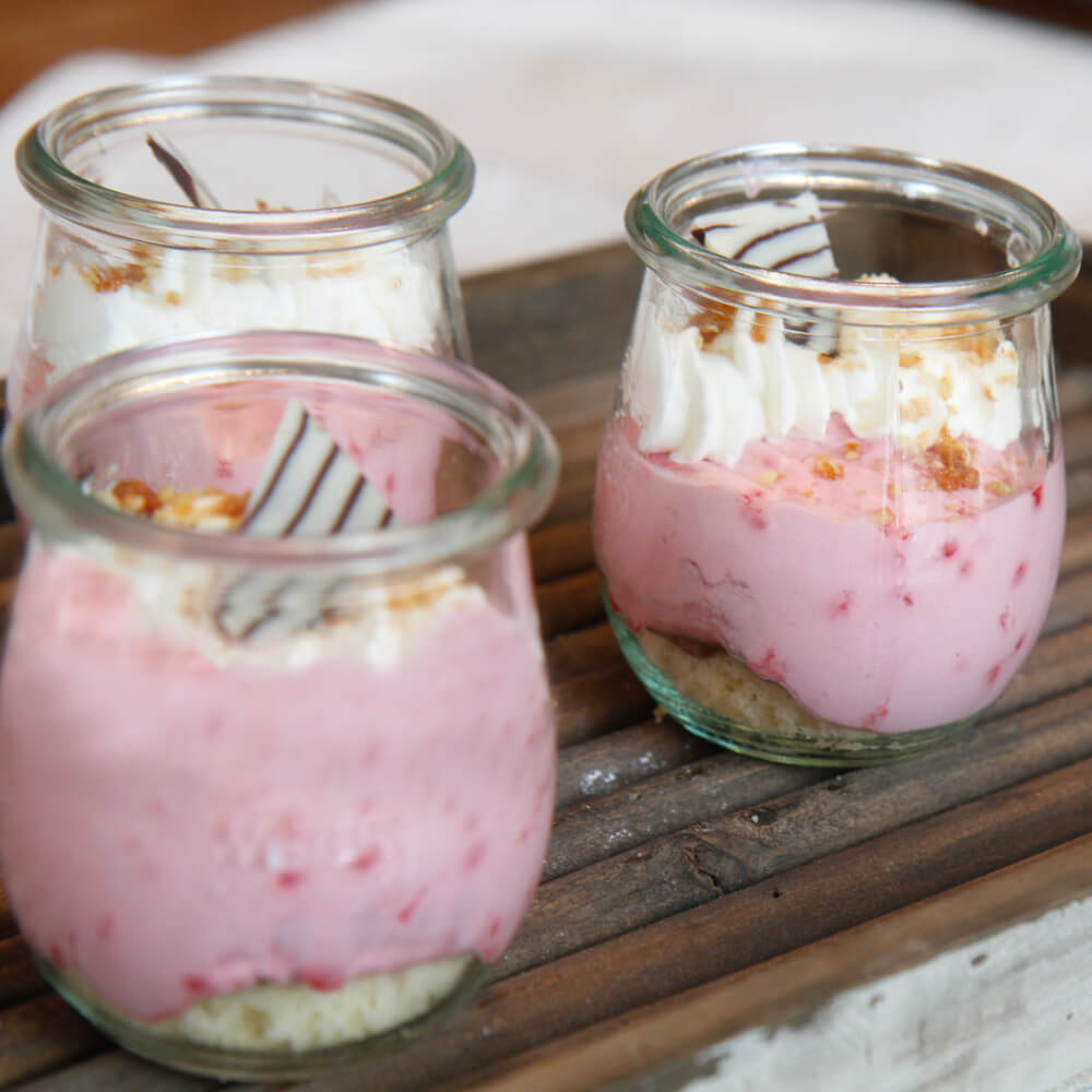 Rezept für Himbeerglas mit Nougatkern 3 Gläser mit Himbeer-Creme, garniert mit Sahne und einem Stück weiß-braun gestreifter Schokolade auf einem Holzbrett serviert