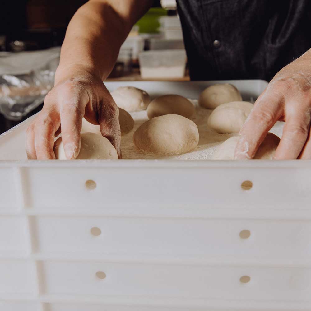 Hand setzt runde Teiglinge in einer weißen Gärbox auf einer bemehlten Arbeitsfläche