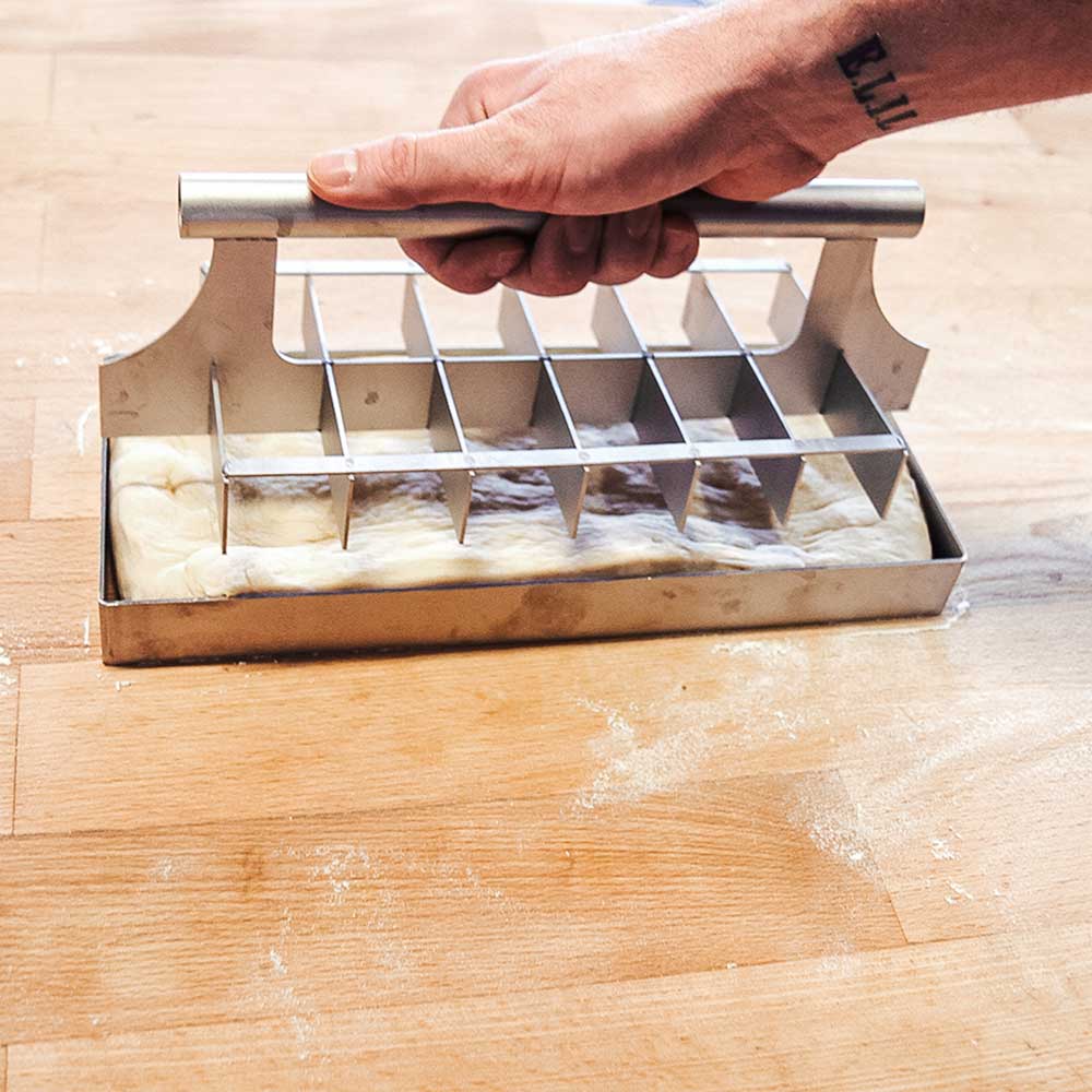 Teigteiler eckig Teigteiler, gefüllt mit Teig. Eine Person drückt das Abstechwerkzeug in den Teig, auf einer Küchenarbeitsplatte.
