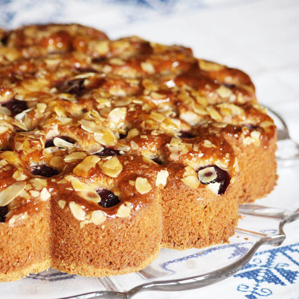 Auf dem Bild befindet sich ein Kuchen in Blumenform. Der Kuchen steht auf einer Glastortenplatte und hat Schokolade und Mandelsplitter als Dekoration