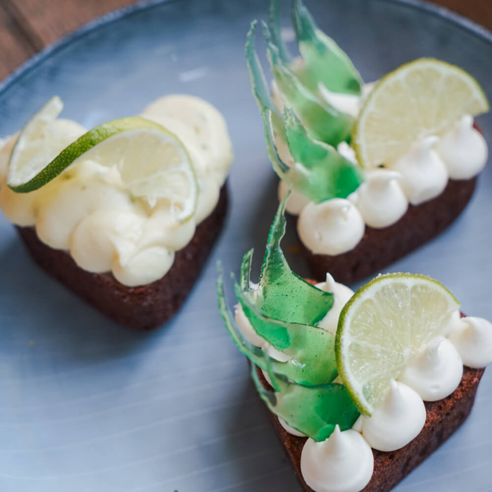 Rezept für Heidelbeer-Limetten-Cupcakes Herzförmige Cupkaes mit einem dunklen Rührkuchenboden mit Heidelbeeren und als Topping eine Sahne-Schoko-Limetten creme. Die Cupcakes sind dekoriert mit einer Limettenscheibe.