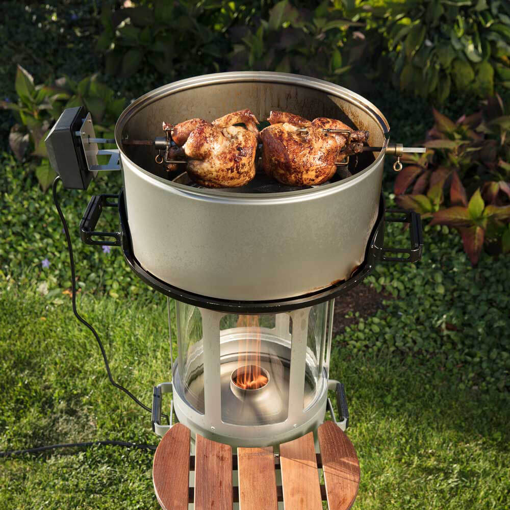 Zwischenring Pelletgrill der angezündet ist und als Grillaufsatz einen Pizzastein mit einem Zwischenring hat. An dem Zwischenring ist ein elektrischer Grillspieß mit 2 Hähnchen befestigt.