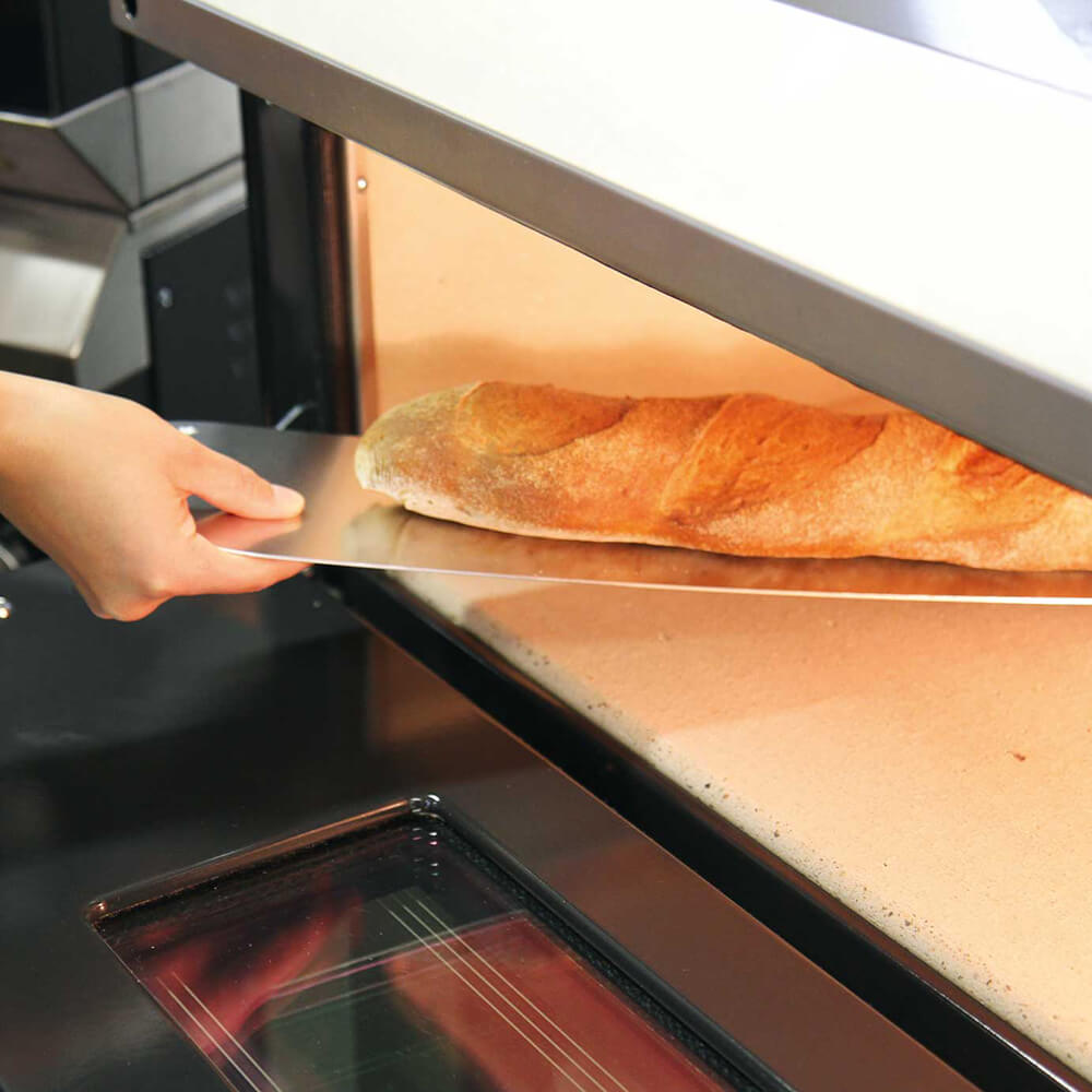 Ein Baguette wird gerade mit dem Baguetteschieber aus Aluminium aus dem Elektroofen geholt