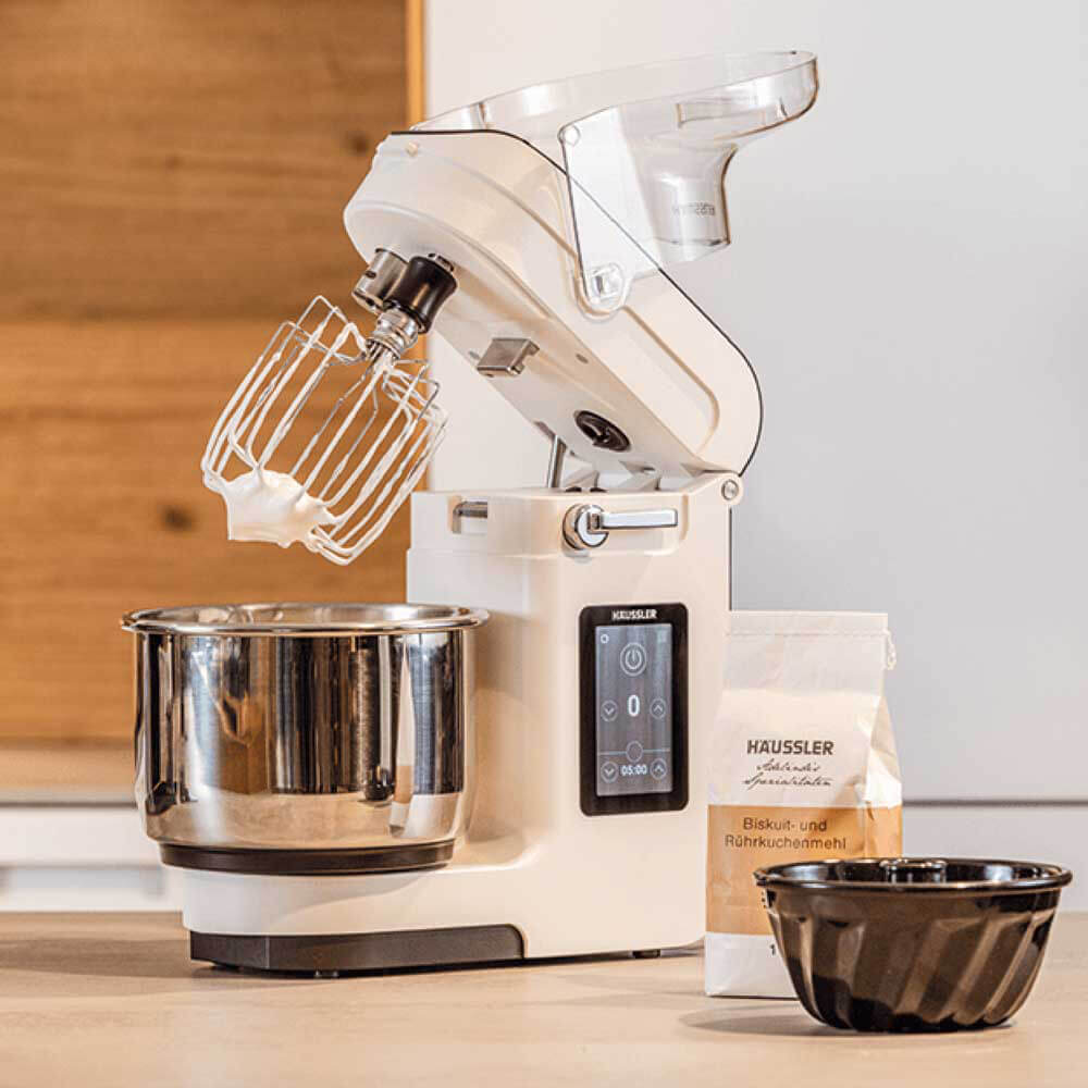 NOVA Teigknetmaschine NOVA Teigknetmaschine mit Rührbeseneinsatz. Am Rührbesen hängt aufgeschlagener Eischnee. Vor der Teigknetmaschine steht eine Backform und ein Mehl auf einer Küchenarbeitsplatte.