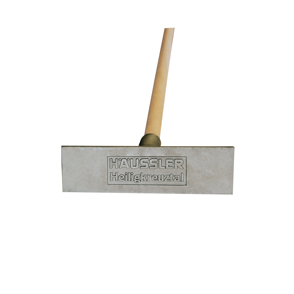 Ascheausziehkrücke Recheckige, aus Aluminiumgussgefertigte Ascheausziehkrücke mit der Prägung "Häussler Heiligkreuztal". Der Stiel ist aus hellem Holz gefertigt.