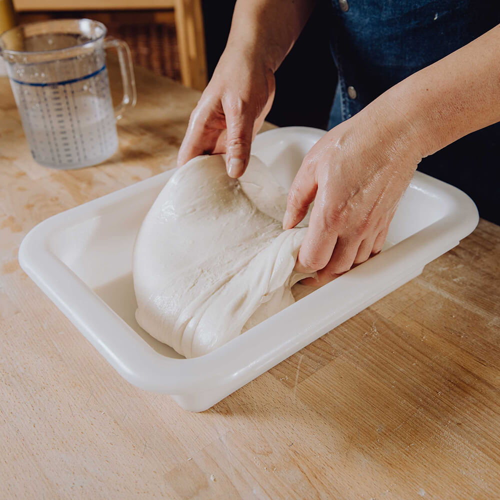 Teigwanne klein Ein Teig wird gerade in eine Teigwanne gelegt. Diese Teigwanne steht auf einer braunen Arbeitsplatte und im Hintergrund befindet sich ein Messbecher mit Wasser