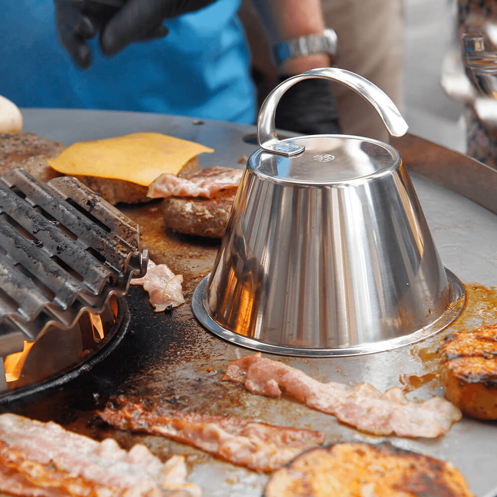 Grill- und Backofenreiniger Es wird eine Häussler Grill Platte mit gebratenem Speck und Fleischpattys gezeigt, in der Mitte der Grillplatte befindet sich eine umgedrehte Edelstahlschüssel mit Griff