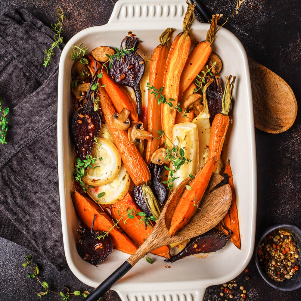 Rezept für Ofengemüse Ofengemüse bestehend aus Karotten, Fenchel, Zwiebeln und frischen Kräutern in einer weißen Auflaufform mit Kochlöffel