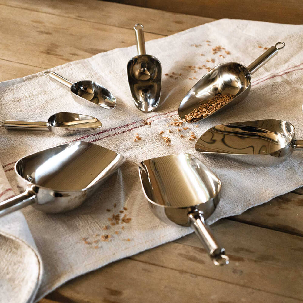 Abwiegeschaufeln aus Edelstahl auf einem Bäckerleintuch im Kreis angeordnet. Einige enthalten verschiedene Getreidesorten. Alles liegt auf einer Holzfläche.