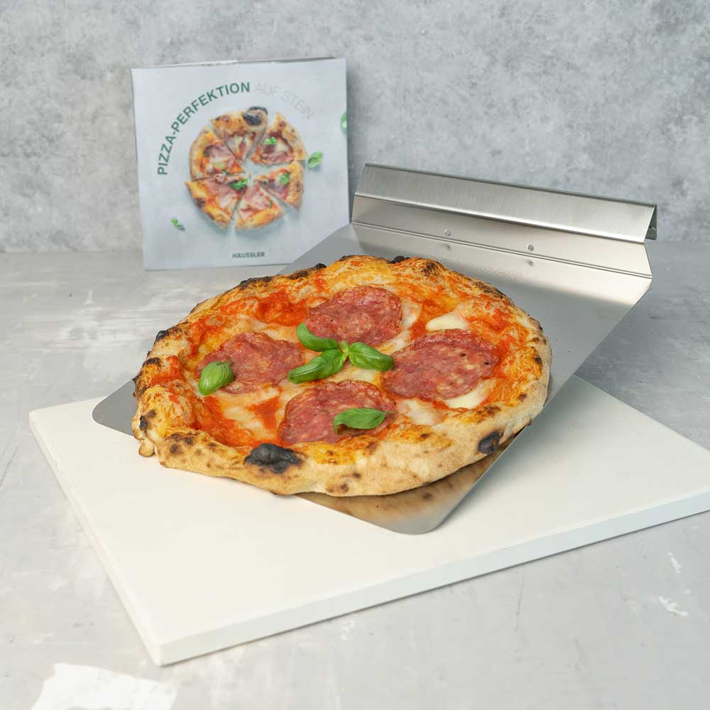 Pizzastein mit Backschieber Weißer Pizzastein, Backschieber mit Handgriff, das Buch Pizza-Perfektion auf Stein und eine Salami-Pizza liegt auf dem Backschieber