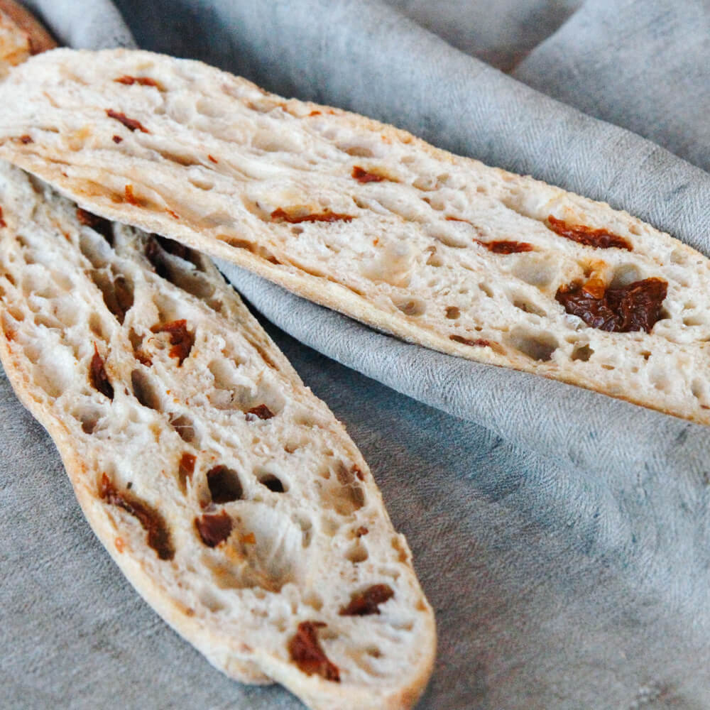 aufgeschnittenes Baguette mit getrockneten Tomaten auf einem grauen Tuch