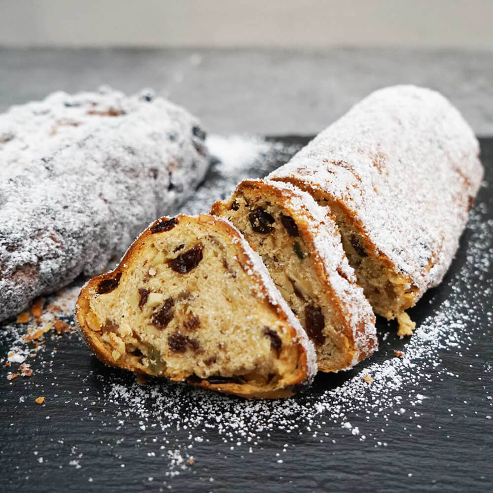 Stollenbackformen mit Deckel Es liegen 2 Christstollen mit Puderzucker bestreut auf einem schwarzen Marmorstein. Ein Christsollen mit Cranberrys und Mandeln ist aufgeschnitten