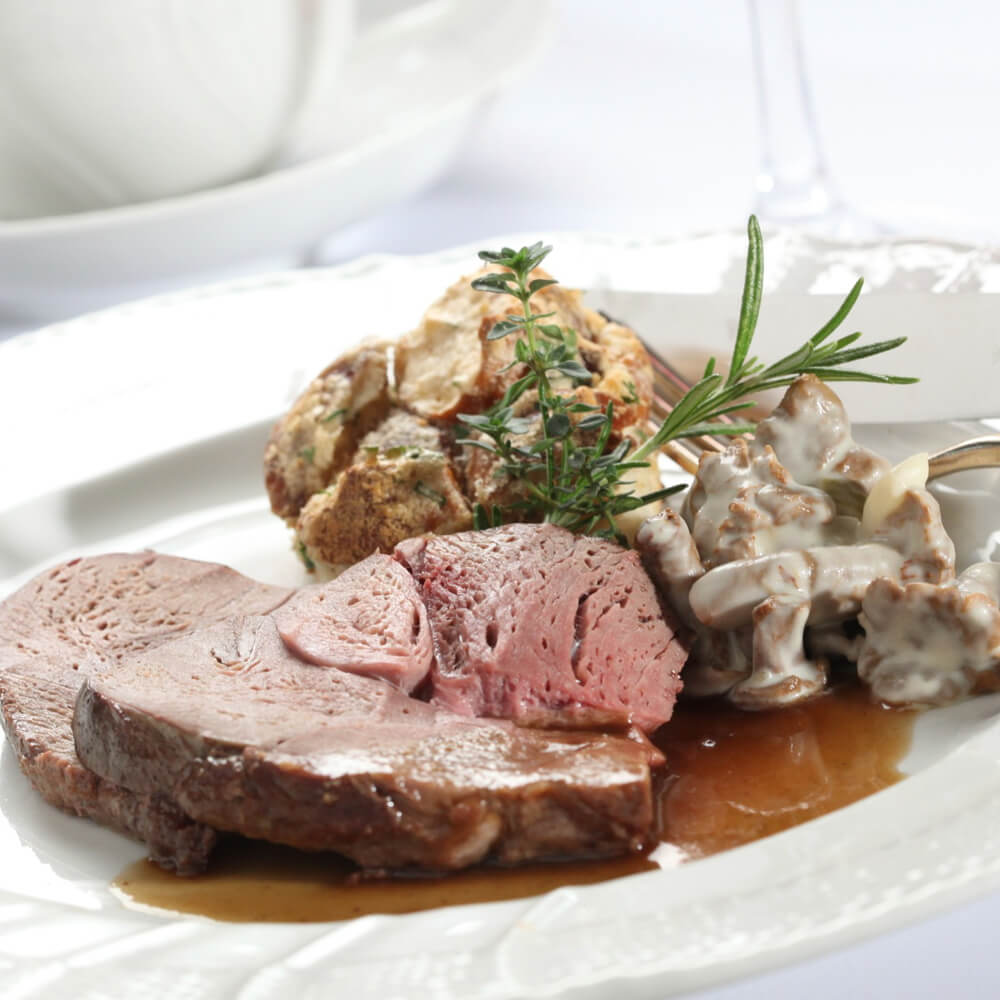 Rezept für Rehkeule mit Rahmpilzen, Rotkraut & Brezelknödelauflauf rosa gebratene Rehkeule mit Pilzen in Rahmsoße und einem Brezelknödel auf einem weißen Teller mit etwas Soße serviert