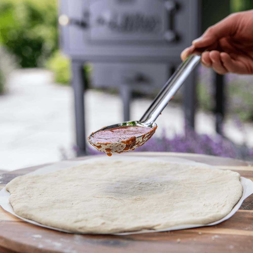 Pizzasugo-Löffel mit Tomatensoße über einem Pizzaboden, im Hintergrund ein Holzofen