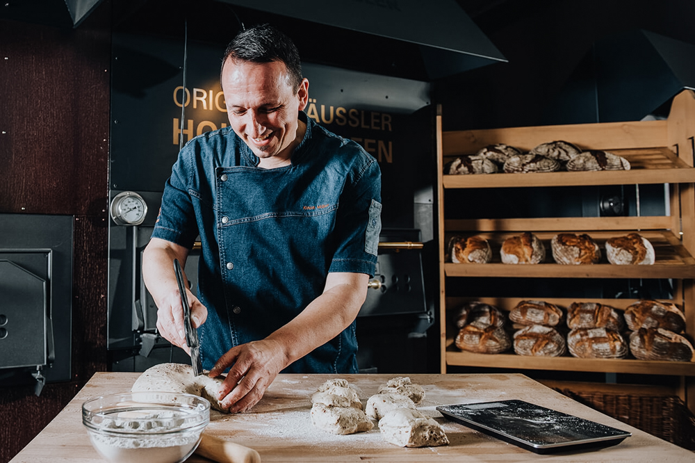 Bäckermeister_in_Backstube Bäckermeister beim händischen Kneten und Teilen von Teig auf einer bemehlten Arbeitsflälche - konzentrierte Handarbeit in traditioneller Backstube vor einem Häussler Holzbackofen.