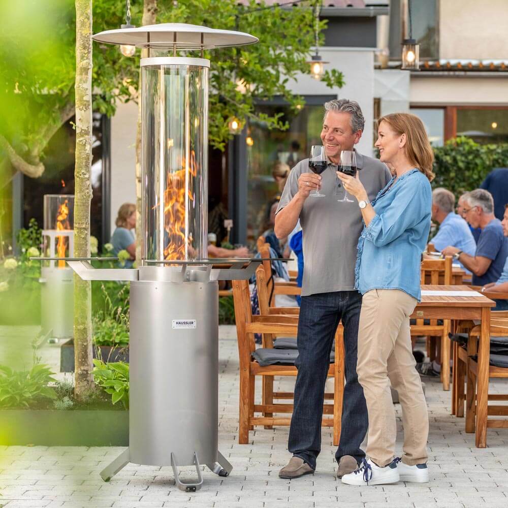 Terrassenstrahler in einem Biergarten. Daneben stehen zwei Personen mit einem Glas Wein und schauen den Terrassenstrahler an.