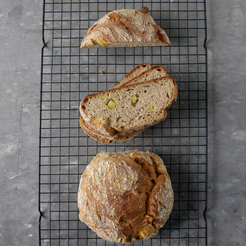 Rezept für Adelindes Würziges Kartoffelbrot 2 Kartoffelbrotlaibe auf einem schwarzen Gitter mit einem dunkelgrauen Hintergrund. Ein Laib ist in Scheiben geschnitten und in den Scheiben sind Kartoffelstücke zu erkennen.