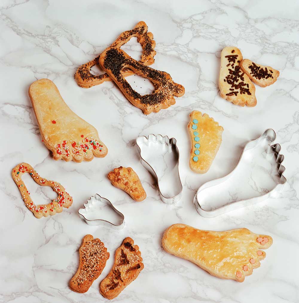 Käsefuß-Ausstecher Drei Fußaustecher in drei verschiedenen Größen. Ringsum liegen Plätzchen mit verschiedenen Dekors auf einer weißen Arbeitsplatte.