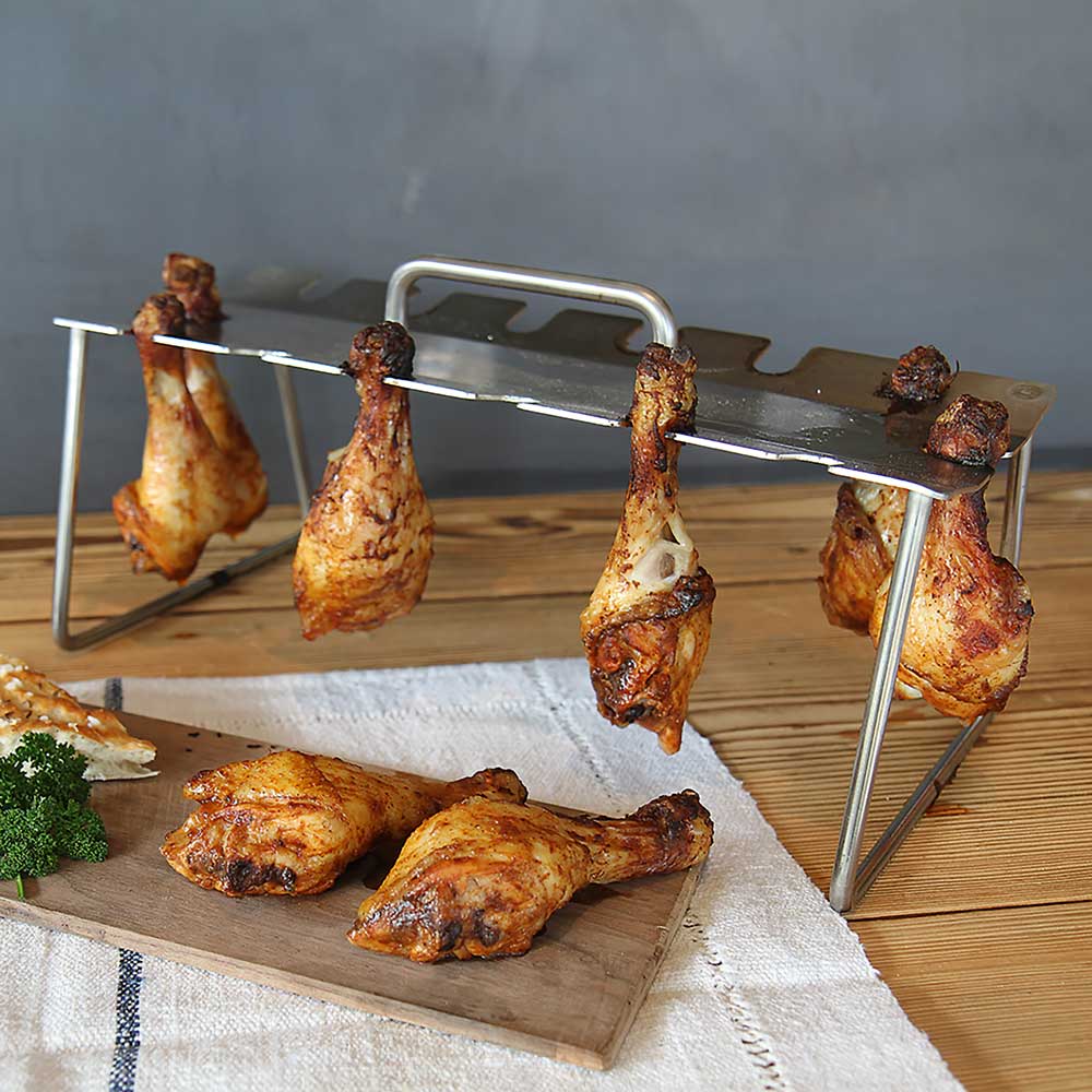 Hähnchenkeulenhalter Hähnchenkeulenhalter der auf einem Tisch steht. Es sind sechs Hähnchenschlegel eingehängt und zwei liegen auf einem Holzbrett davor.