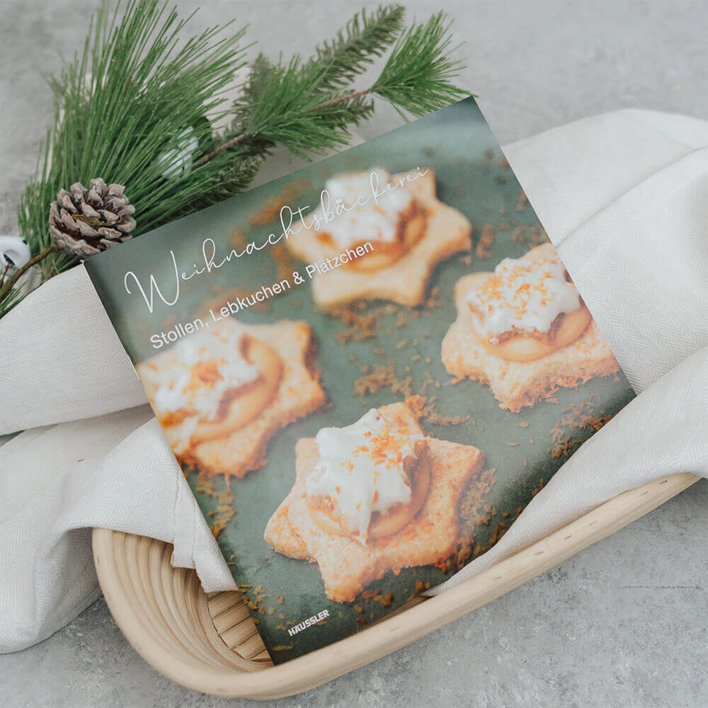 Das Häussler-Buch "Weihnachtsbäckerei" liegt in einem mit einem beige-farbenen Leinentuch gefüllten Gärkörbchen. Daneben sind ein paar grüne Zweige. 