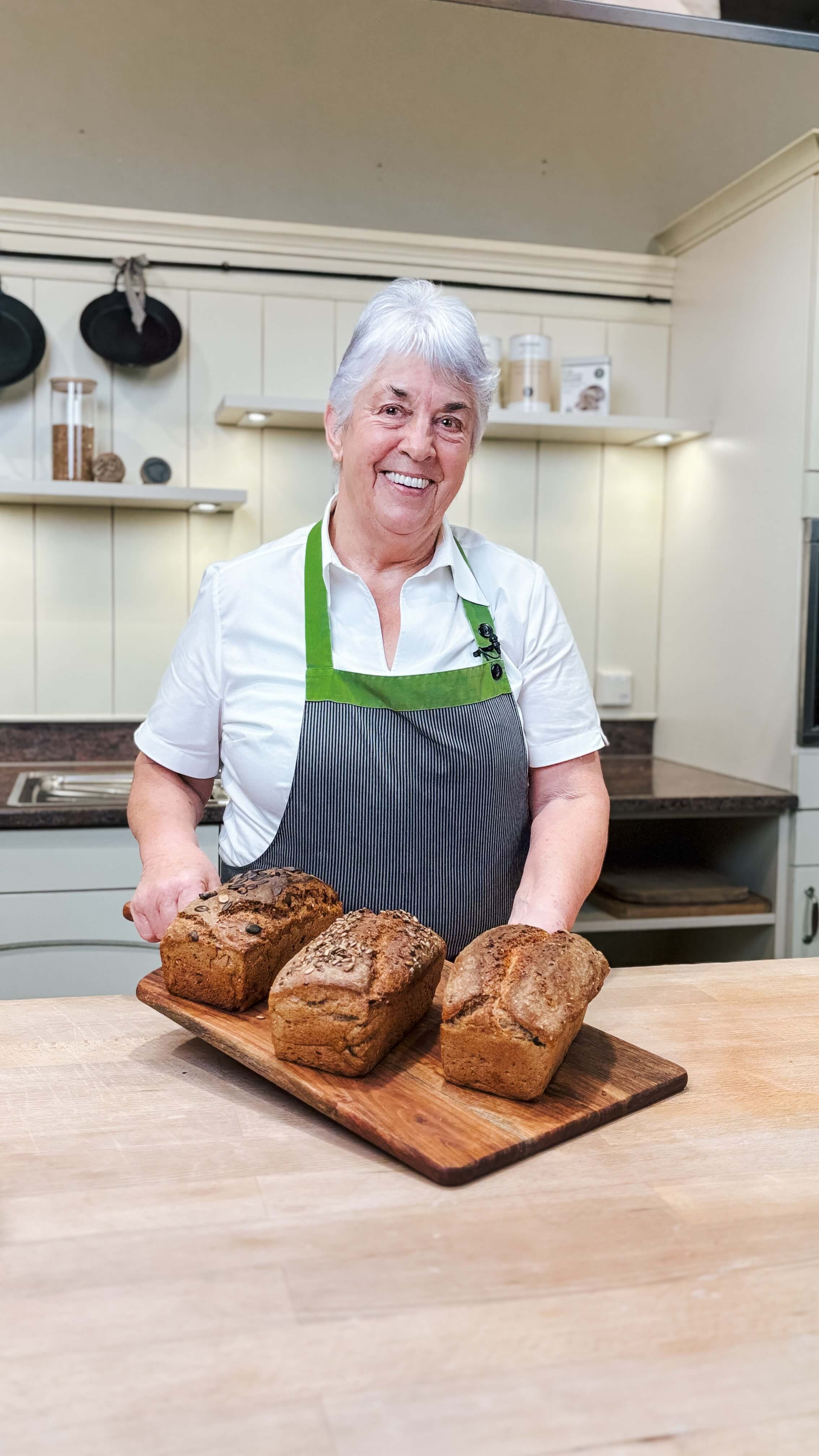 Rezept für Adelindes traditionelles Dinkel-Vollkornbrot Adelinde Häussler trägt eine grau gestreifte Schürze und hält einen Backschieber in der Hand auf dem Ihre drei Selbstgebackenen Dinkelbrote liegen
