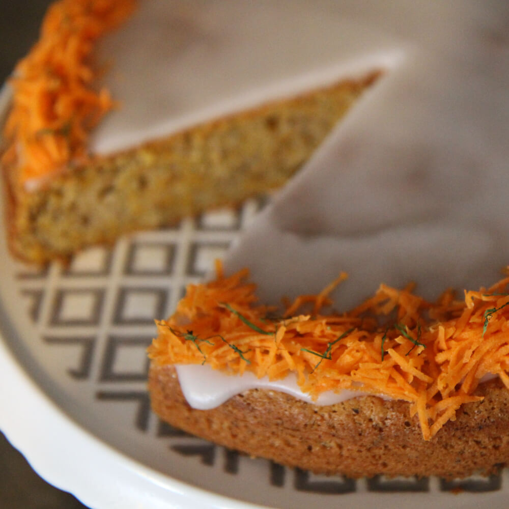 Rezept für Rüblikuchen saftiger Rüblikuchen dekoriert mit Zuckerguss und frisch gerapselten Karotten auf einer weiß-grau karierten Kuchenplatte