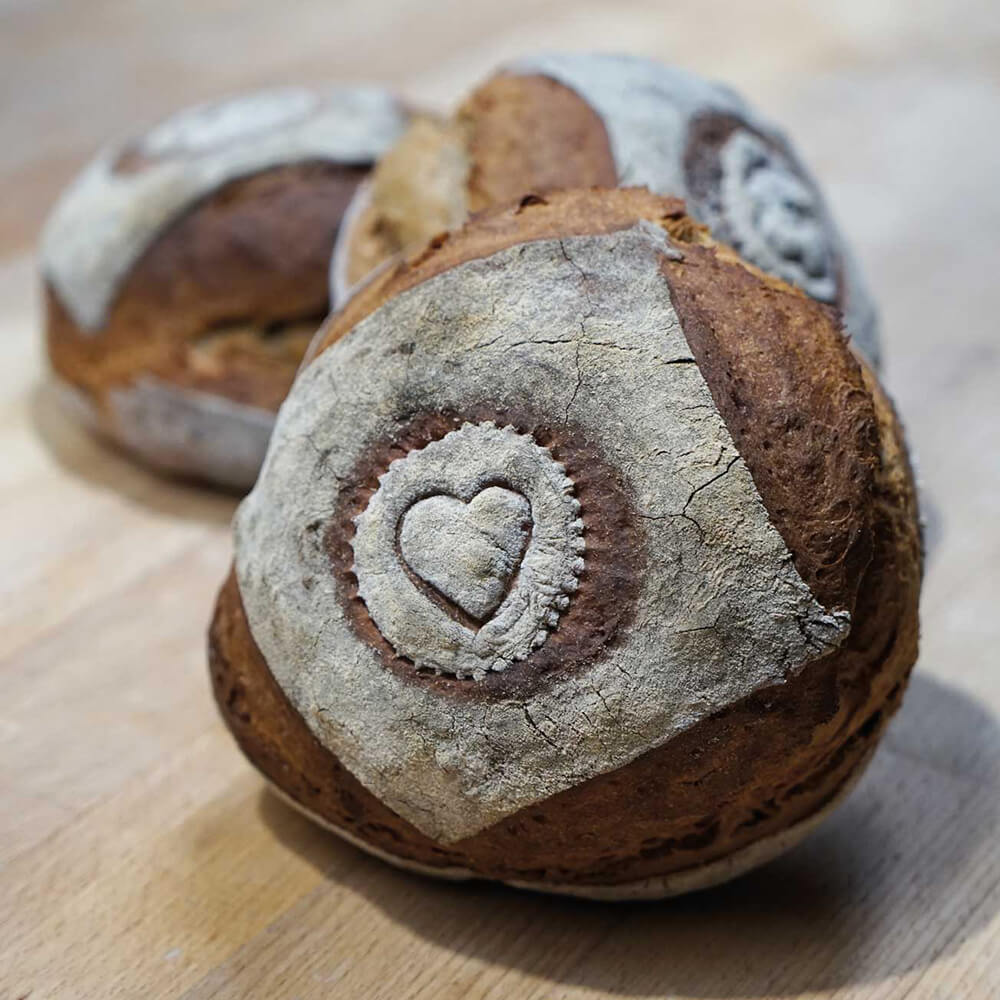 3 Brotlaibe auf einem Tisch. Der vorderste Laib hat ein Herz-Motiv in der Mitte. 