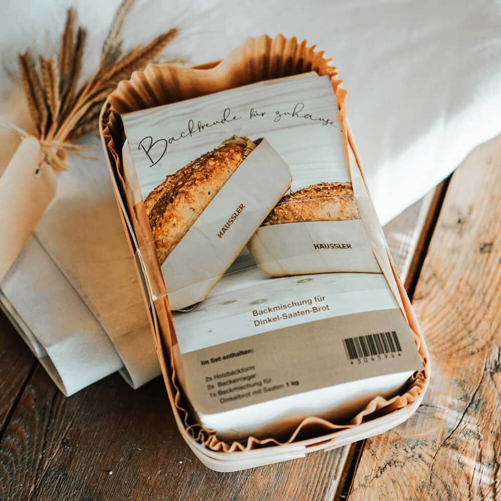 Holzbackform-Set von Häussler mit Backmischung für Dinkel-Saaten-Brot, verpackt in Papier, auf einem Holztisch neben einem Leinentuch und dekorativen Getreidehalmen.