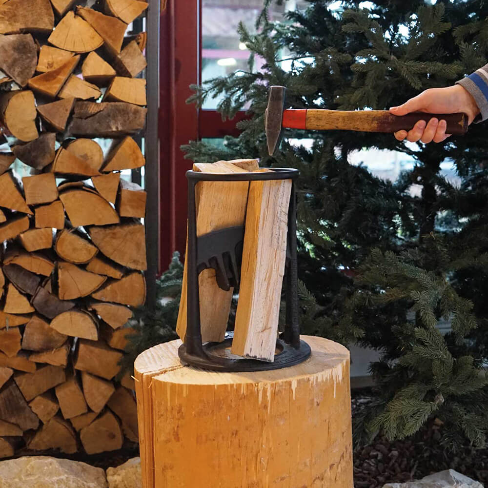 Holzspalter Manueller Holzspalter auf Baumstumpf, Person spaltet Holz mit Hammer, gestapeltes Holz und Nadelbaum im Hintergrund.