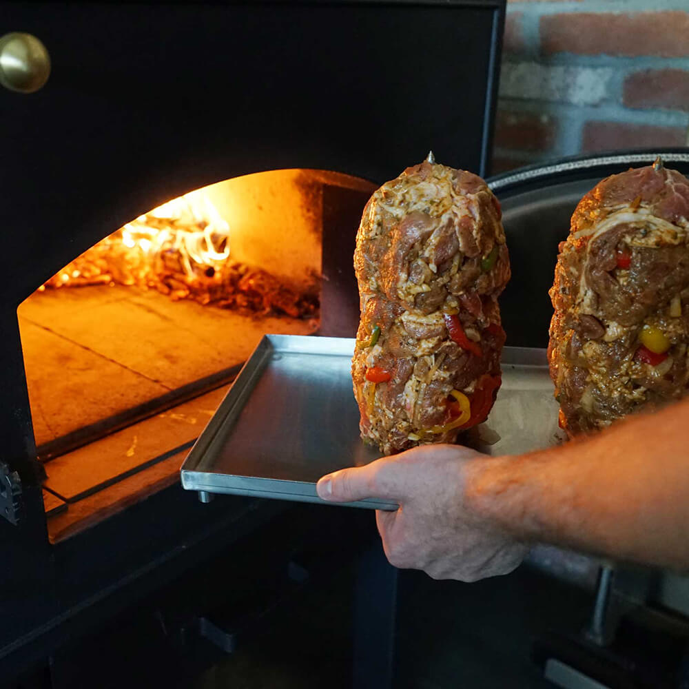 Gyrosspieß 38 x 23 x22 cm Fleisch und Gemüse auf zwei Gyrosspieße geschichtet, die gerade in den Holzbackofen geschichtet werden, im Hintergrund brennt im Ofen eine Flamme