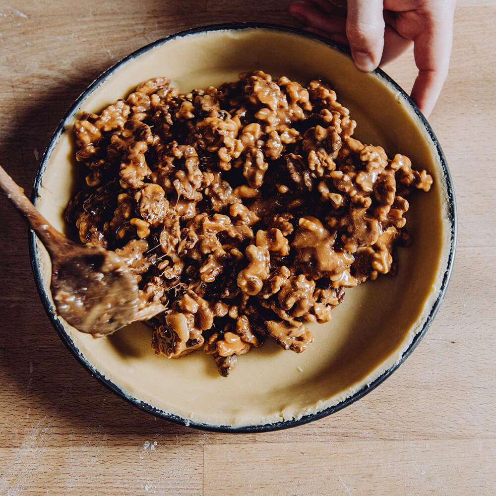 Blauglanzrundform Eine Blauglanzrundform die innen mit Teig ausgekleidet ist, wird mit einer Füllung gefüllt. Die Form steht auf einem braunen Arbeitstisch