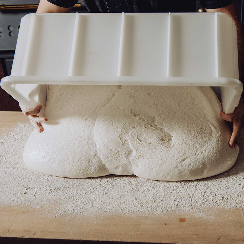 Eine Teigwanne mit Teig wird gerade umgedreht, sodass der Teig auf eine bemehlte Arbeitsfläche fällt
