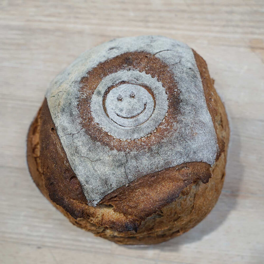 Ein Brotlaib liegt auf einem Holztisch. In der Mitte des Laibs ist ein Smiley-Motiv aufgedrückt.
