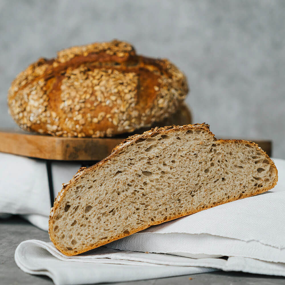 Rezept für Dinkel-Älbler Brot frisch gebackenes Brot mit Dinkelflocken bestreut. im Vordergrund ist ein halbes und im Hintergrund ein ganzes Brot zu sehen vor einem hellgrauen Hintergrund.
