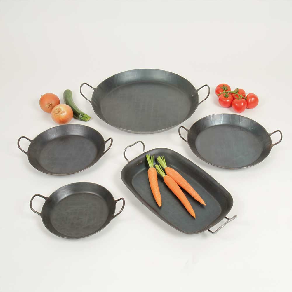 Fünf Eisenpfannen in verschiedenen Größen mit zwei Griffen. Vier Pfannen sind rund eine Pfanne ist Rechteckig. In der Recheckigen Pfanne liegen Karotten. Außerdem sind Tomaten, Zwiebeln und eine Zucchini zu sehen. 