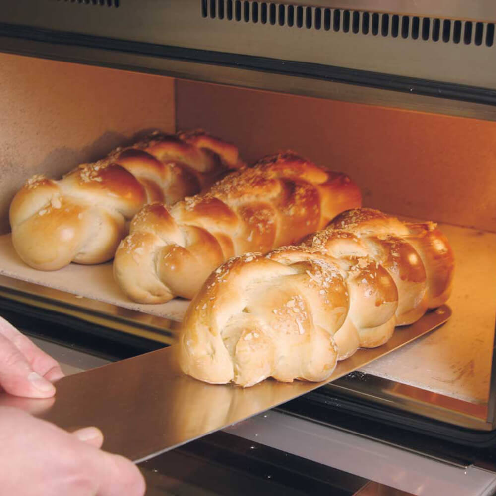 Baguetteschieber 3 Hefezöpfe mit Mandelsplitter liegen nebeneinander in einem Backofen und eines davon wird gerade mit einem Baguetteschieber aus dem Ofen geholt