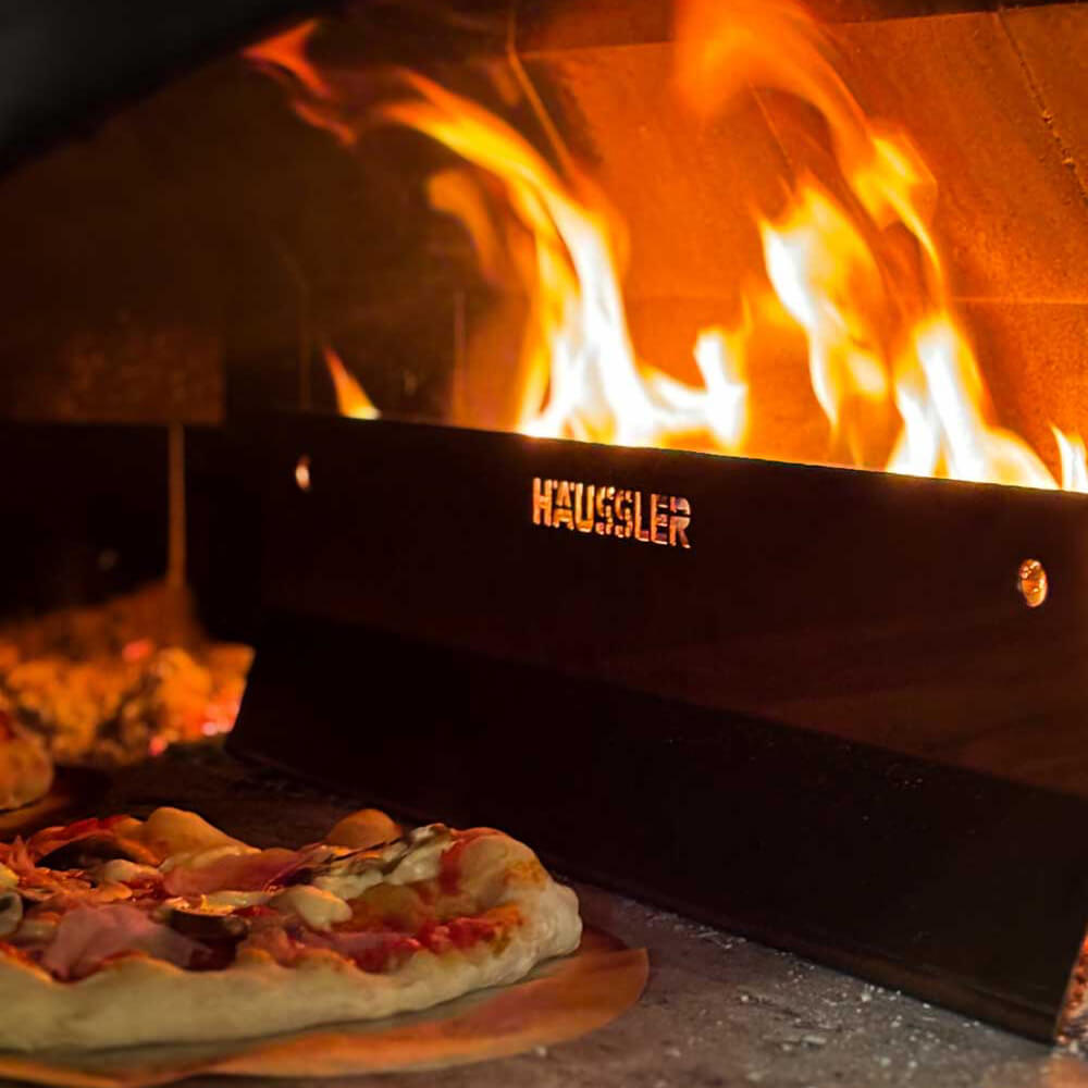 Prallblech für den Holzbackofen Links im Ofen liegt eine Pizza auf dem Stein. Daneben das Prallblech, um die Pizza vor dem Feuer zu schützen