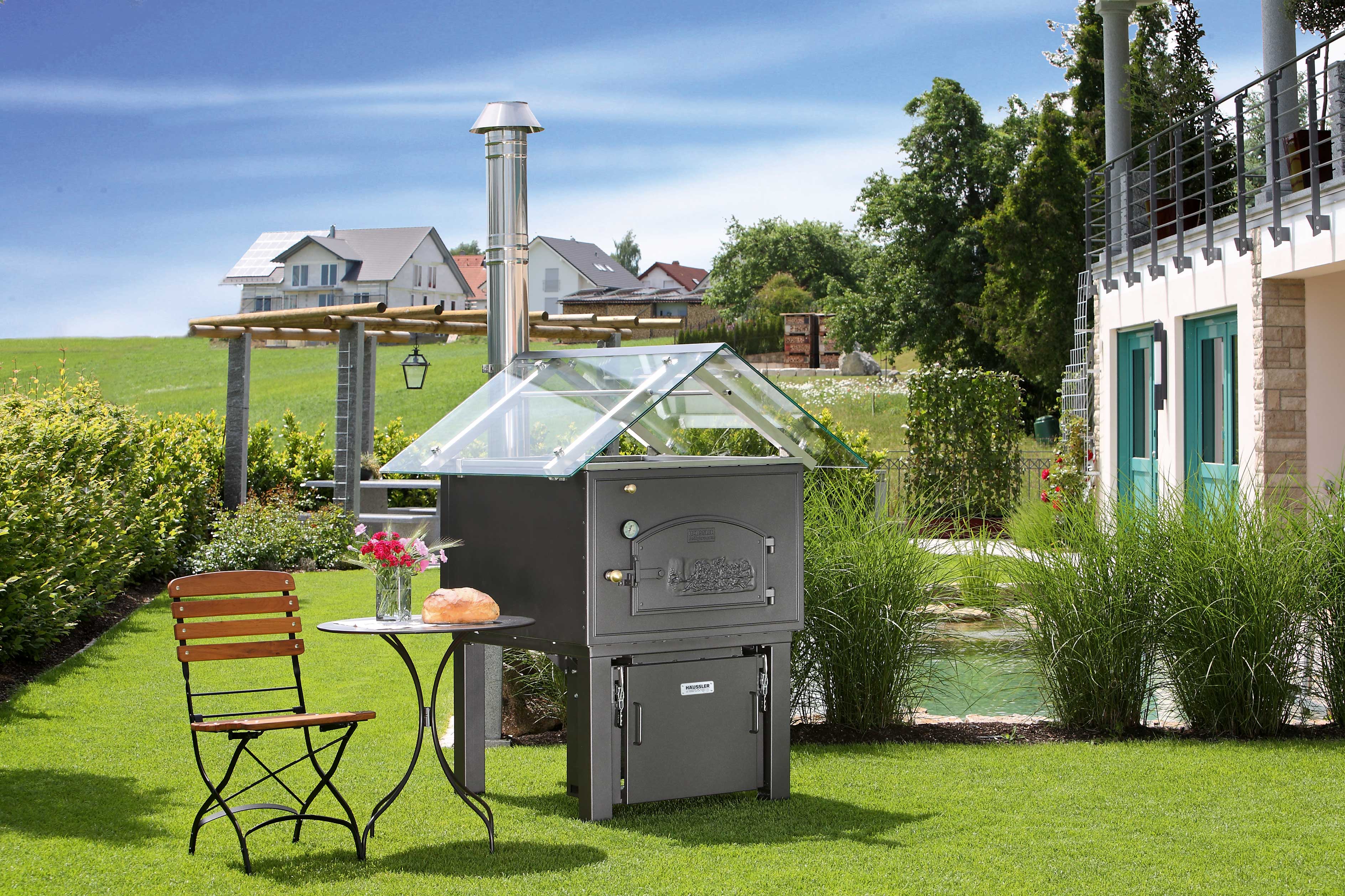 Brennerkassette für den Holzbackofen Holzbackofen mit Brennerkassette, Glasdach und einem Edelstahl-Rauchrohr in einem Garten. Im Hintergrund sind andere Häuser zu sehen und ein Teich. Links neben dem Ofen ist ein Klappstuhl und ein kleiner Tisch aufgebaut.