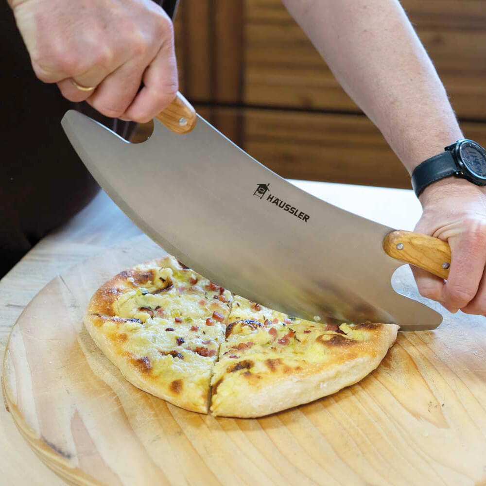 Pizza-Wiegemesser in der Anwendung beim Schneiden einer frisch gebackenen Dinnete auf einer Holzplatte.