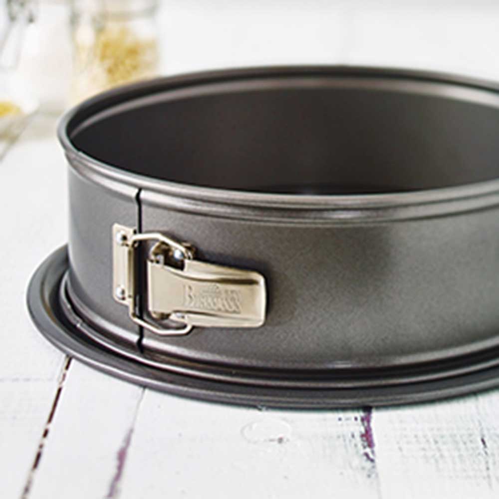 Eine Graue Springform mit Boden und Edelstahl-Verschluss steht auf einem Hellgrauen Holztisch