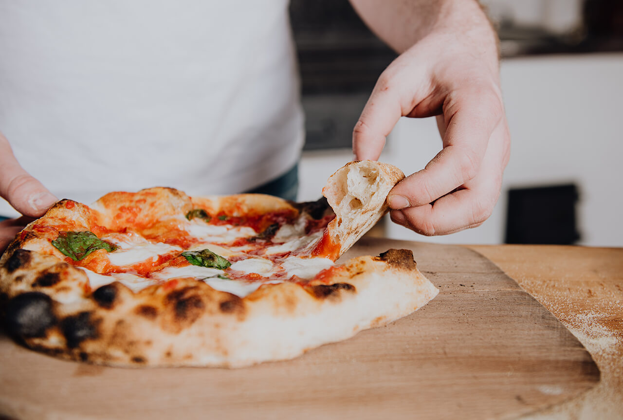 Pizza_auf_Holzbrett Frische Pizza mit knusprigem Rand, angeschnitten und auf einem Holzbrett serviert – der geschmolzene Käse und Belag sind gut sichtbar.
