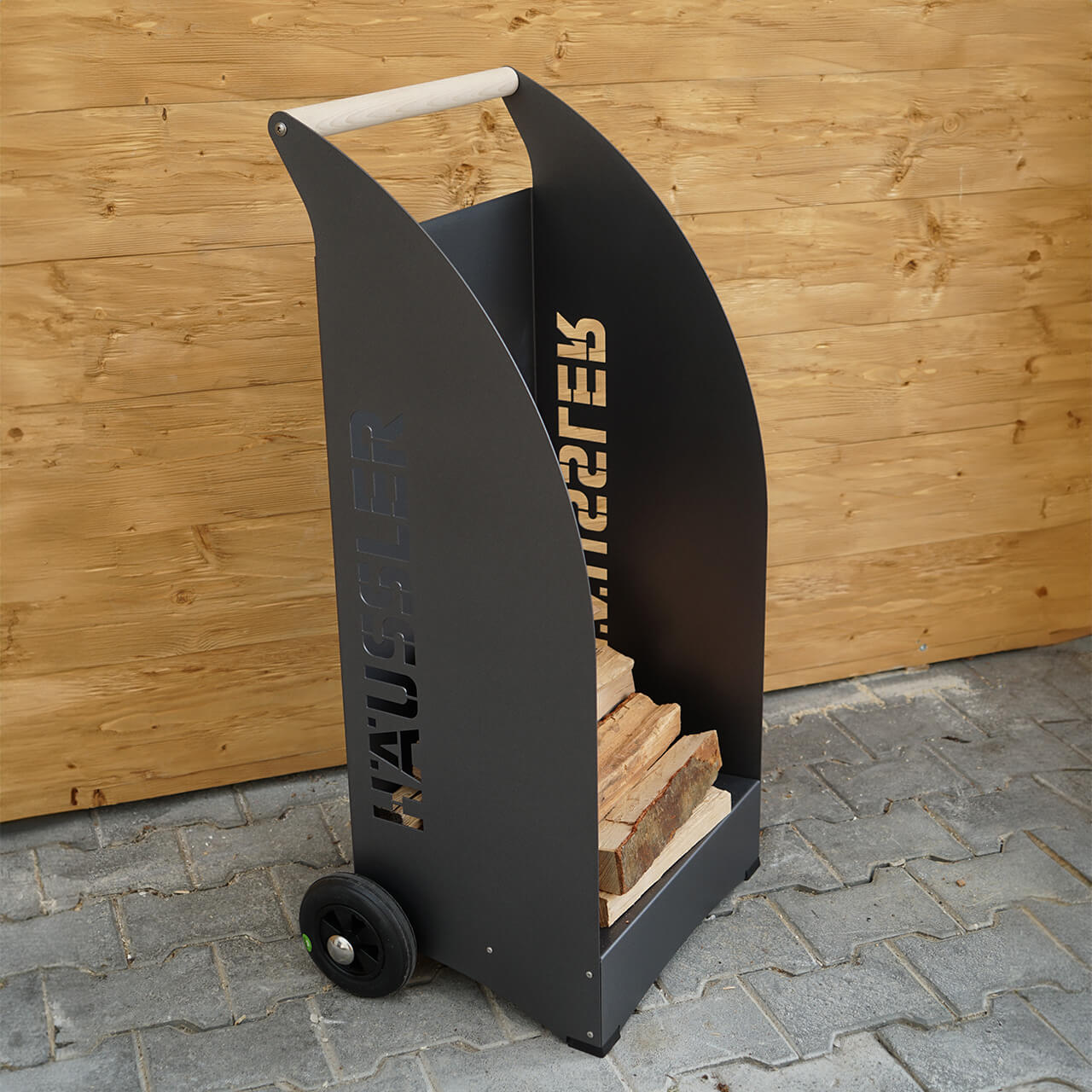 Häussler_Holzwagen Kleiner Holzwagen zur Aufbewahrung von Holzscheiten aus dunklem Metall mit dem Häussler Logo an der Seite. Ideal für Holzbackofen oder offene Kamine.
