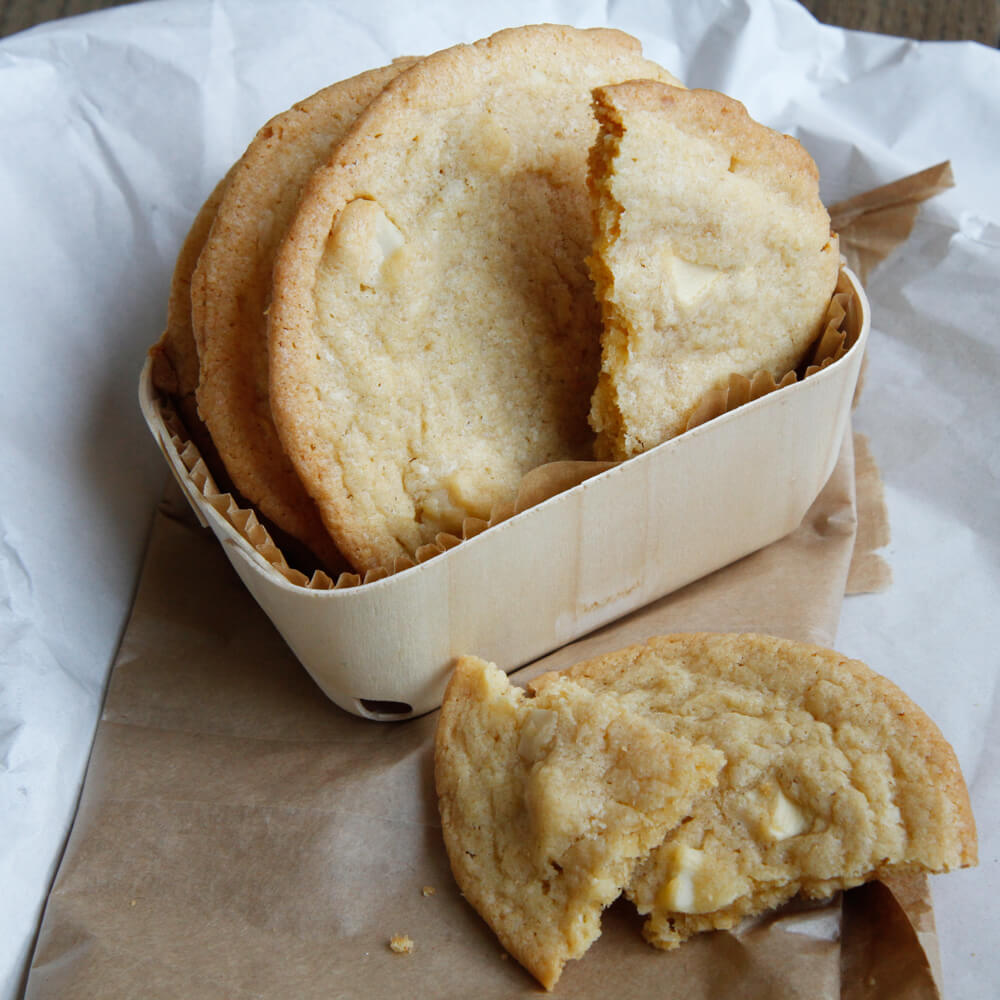 Rezept für White Chocolate Macadamia Cookie selbstgemachte Cookies mit Macadamia und weißer Schokolade in einer Holzbackform serviert. Im Vordergrund ist ein auseinander gebrochener Cookie zu sehen