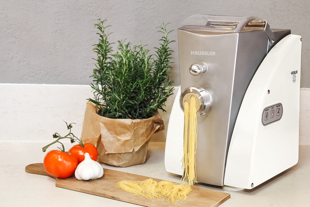 Nudelmaschine Luna mit frischer Pasta auf dem Holzbrett, dekoriert mit italienischen Elementen wie Tomaten, Knoblauch und Rosmarin.