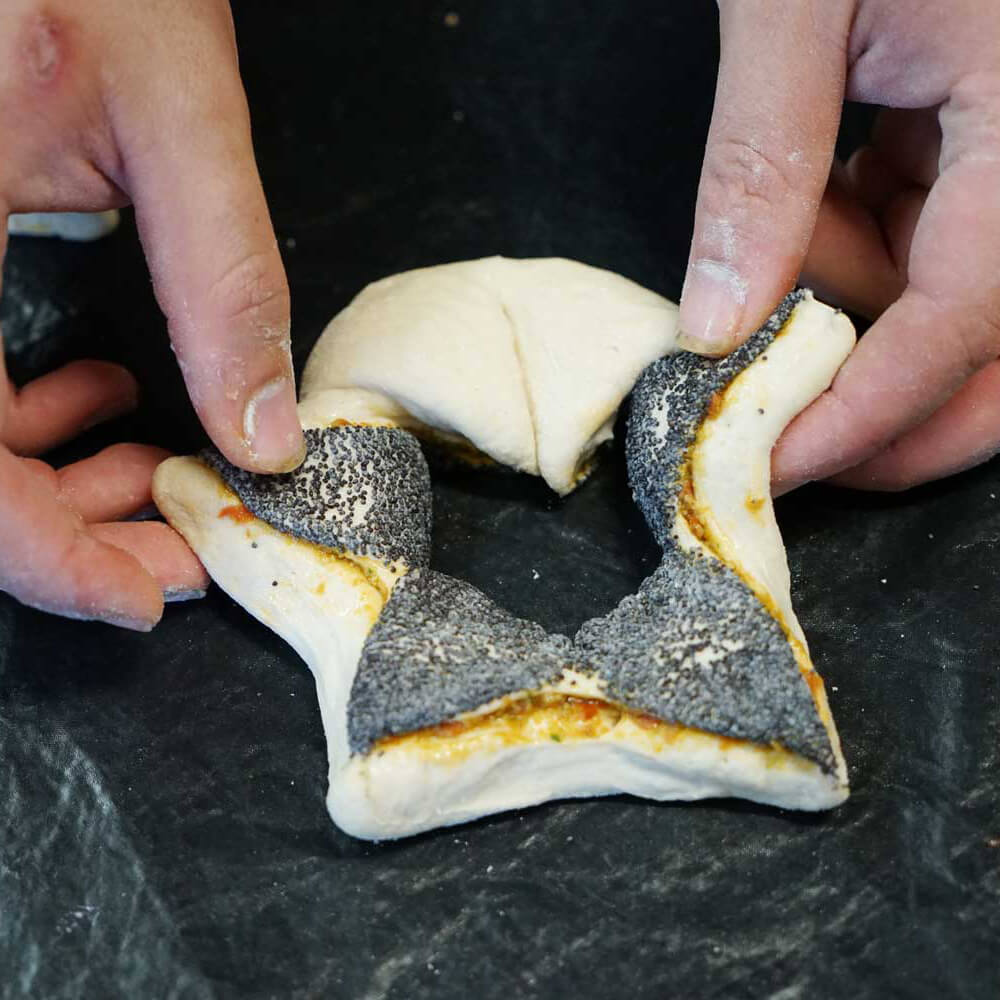 Der Teigling, der eine Sternformartige Form hat sowie ein Loch in der Mitte wird auf eine schwarze Folie gelegt. Auf dem Sternformartigen Brot ist Mohn gestreut. 