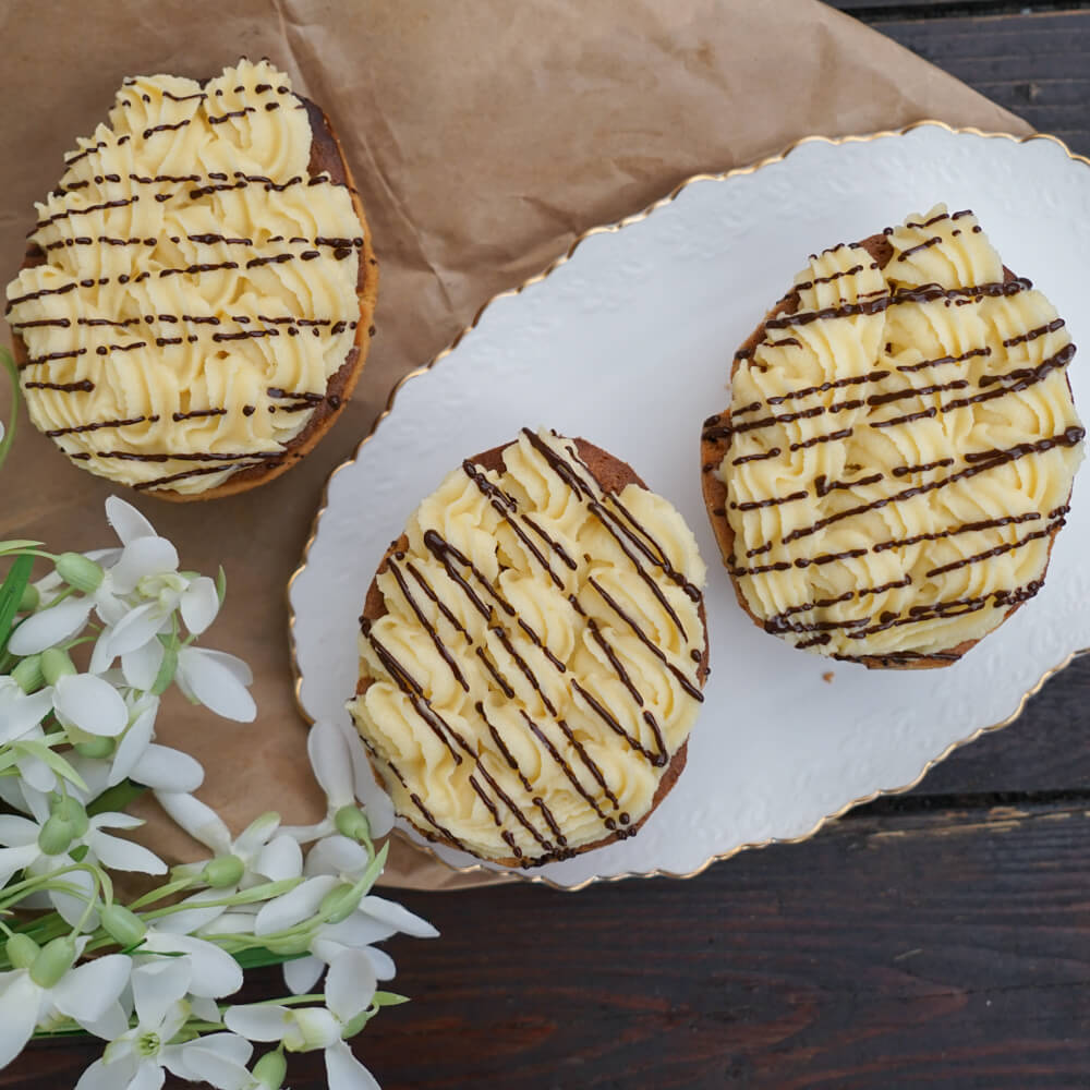Rezept für Eierlikör-Eier Eierlikör-Eier aus Biskuit und einem Sahne-Eierlikörtopping, verziert mit flüssiger Schokolade auf einer ovalen, weißen Kuchenplatte mit ein paar weißen Blumen im Vordergrund