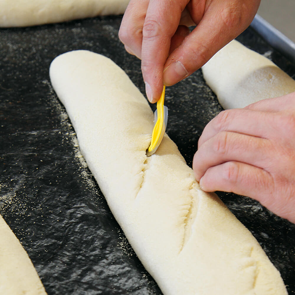 Teigritzmesser Ein Baguette Teigling auf einer Dauerbackfolie liegend, dass mit einem kleinen gelben Messer mit Rasierklinge eingeschnitten wird