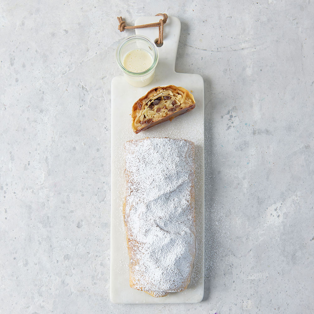 selbstgemachter Apfelstrudel bestreut mit Puderzucker auf einem Brett, mit einem Glas Vanillesoße serviert